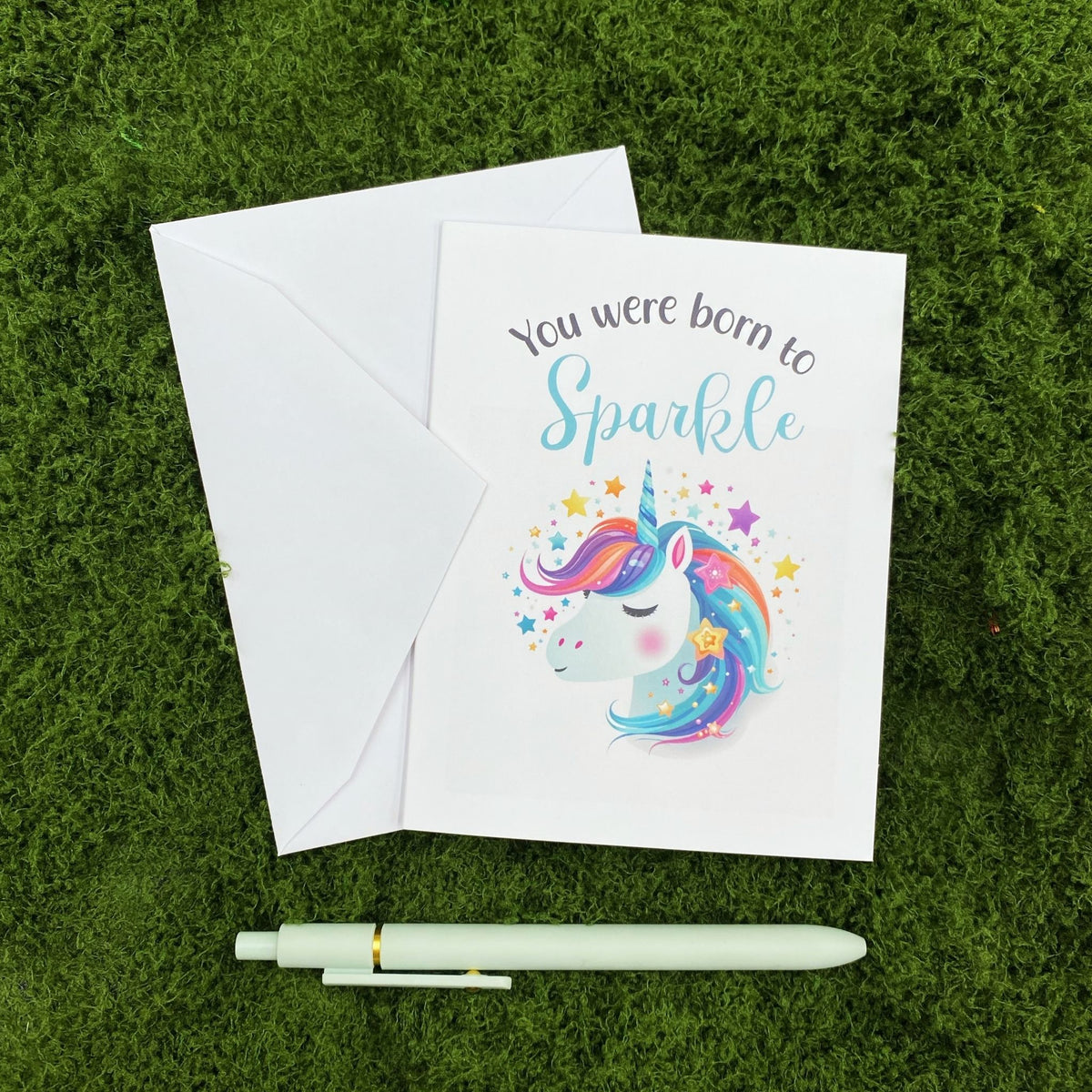 "You Were Born to Sparkle" greeting card lying on a white envelope on grass, with a mint green pen beside it for scale.