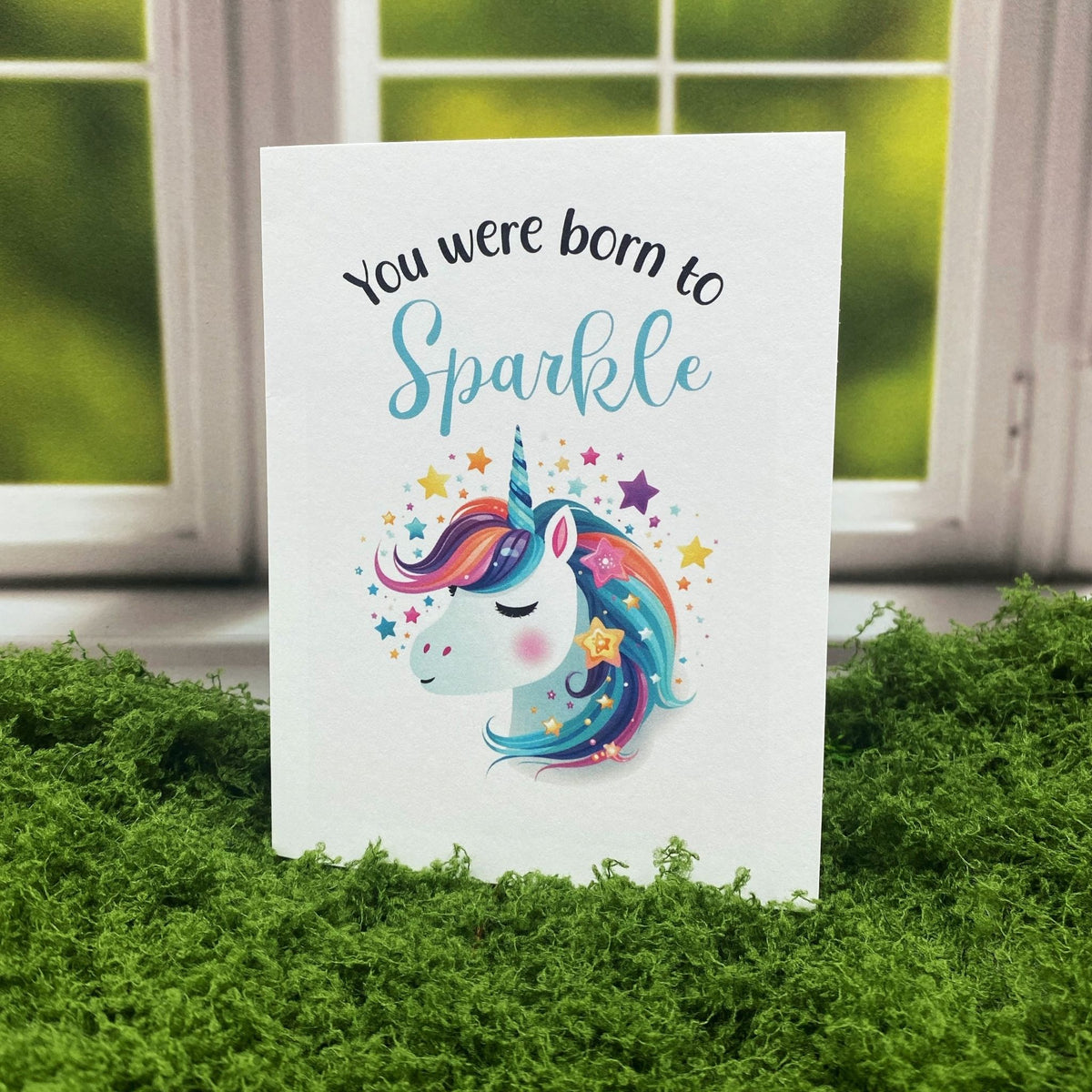 "You Were Born to Sparkle" greeting card standing upright in front of a window, featuring an illustrated unicorn with a rainbow mane surrounded by colorful stars.