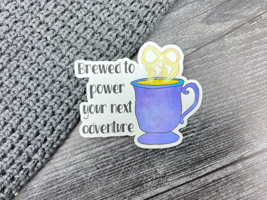Die-cut vinyl sticker showing a steaming purple mug with an infinity symbol in the steam, with the text "Brewed to power your next adventure," on a gray knit and wood background