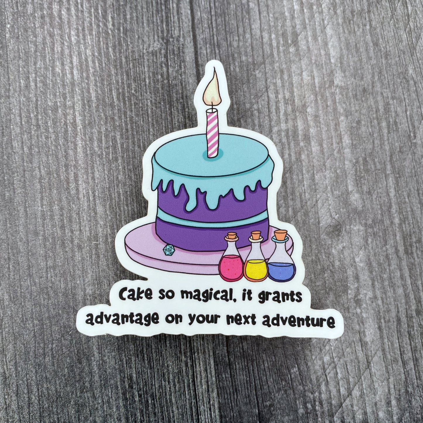 Die-cut vinyl sticker showing a birthday cake with a lit candle next to three colorful potion bottles and a d20, with the text "Cake so magical, it grants advantage on your next adventure," on a gray wood background