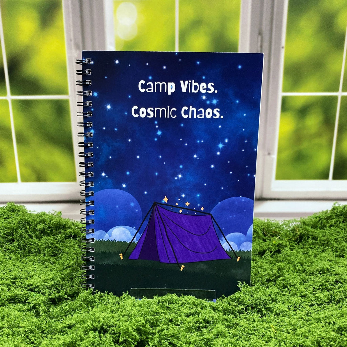 Spiral-bound notebook with a starry night sky illustration and a glowing purple tent, reading 'Camp Vibes. Cosmic Chaos.,' displayed on a moss mat with a window backdrop