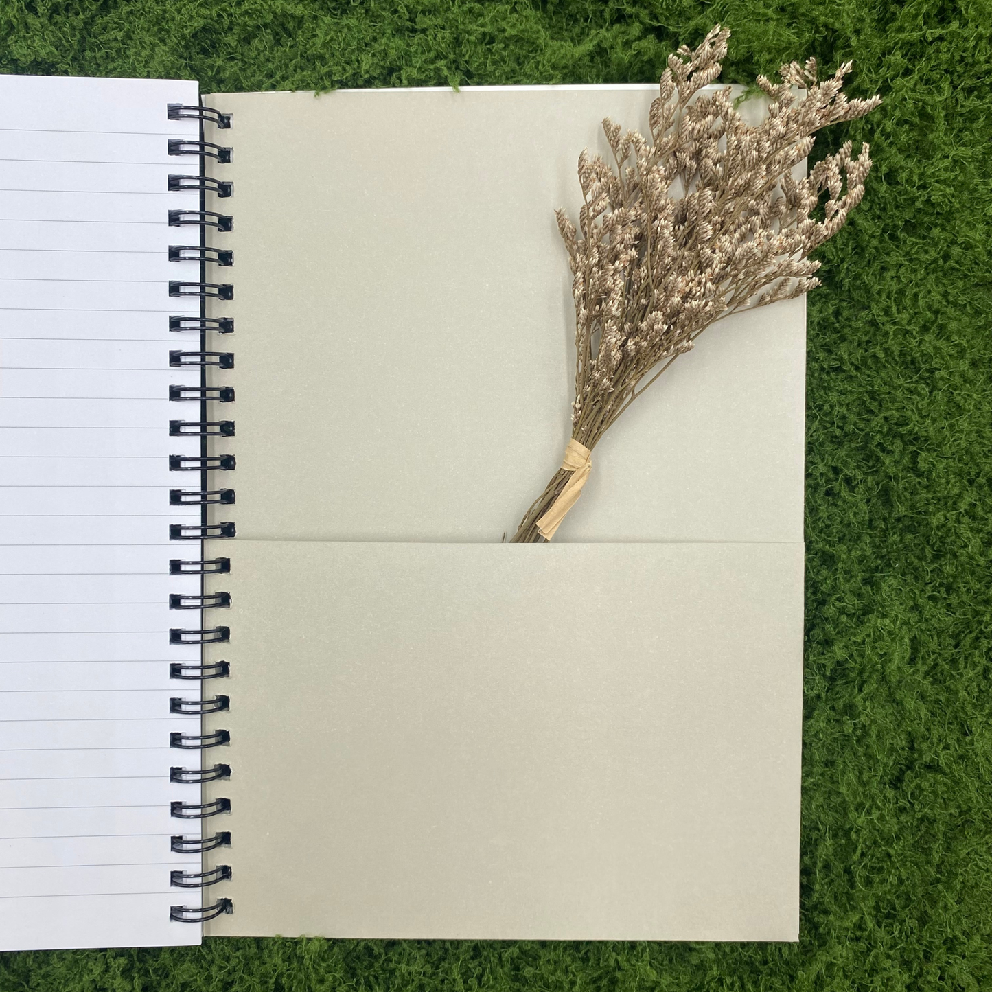 Tan document pocket inside the back cover of a spiral notebook, shown with a bundle of dried flowers tucked partway inside, on artificial grass.