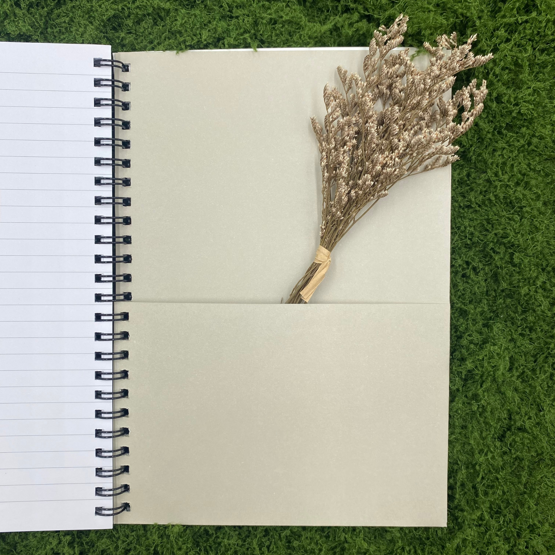 Tan document pocket inside the back cover of a spiral notebook, shown with a bundle of dried flowers tucked partway inside, on artificial grass.