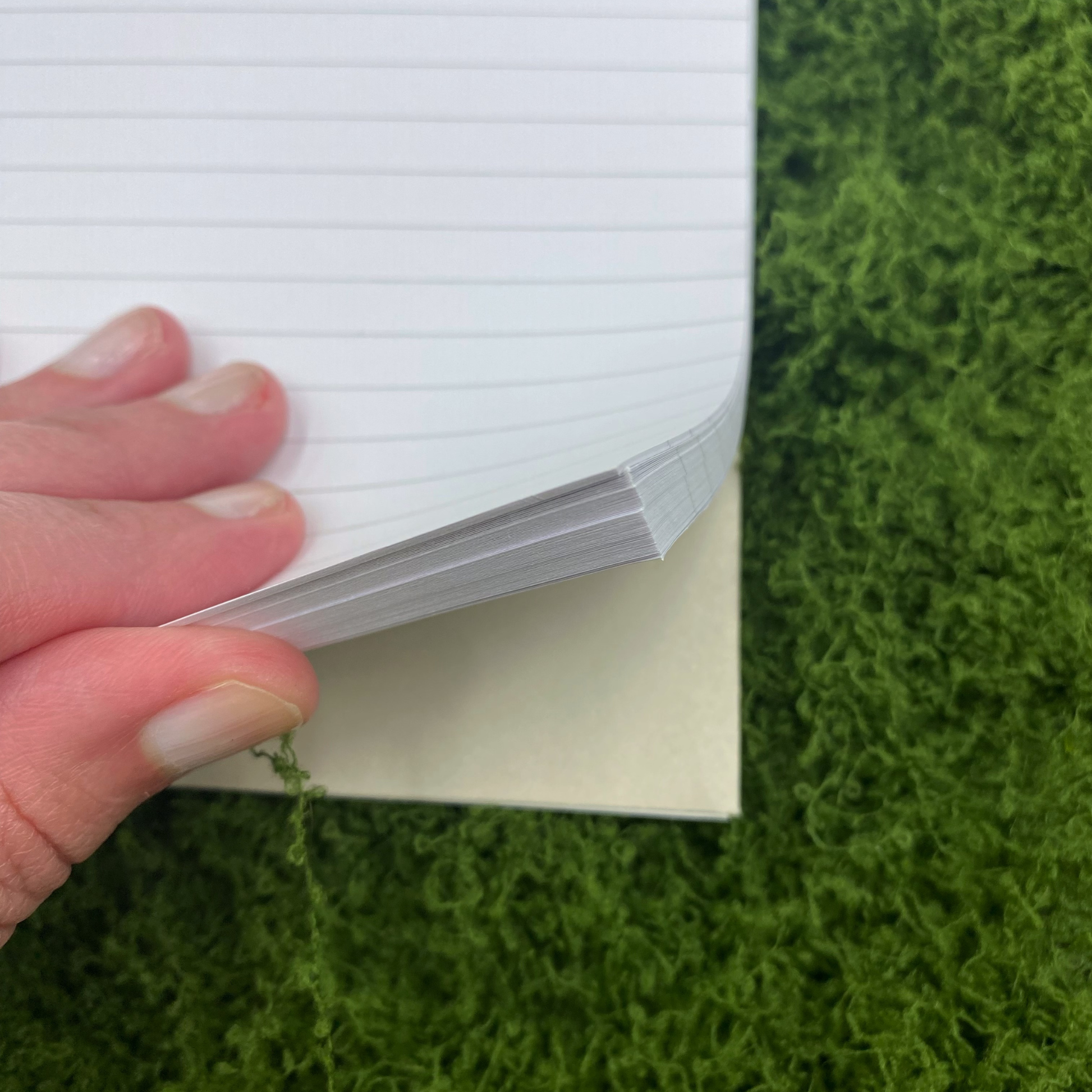 Close-up of lined notebook pages being flipped by hand, showing the paper stack and cream-colored back cover, on artificial grass.