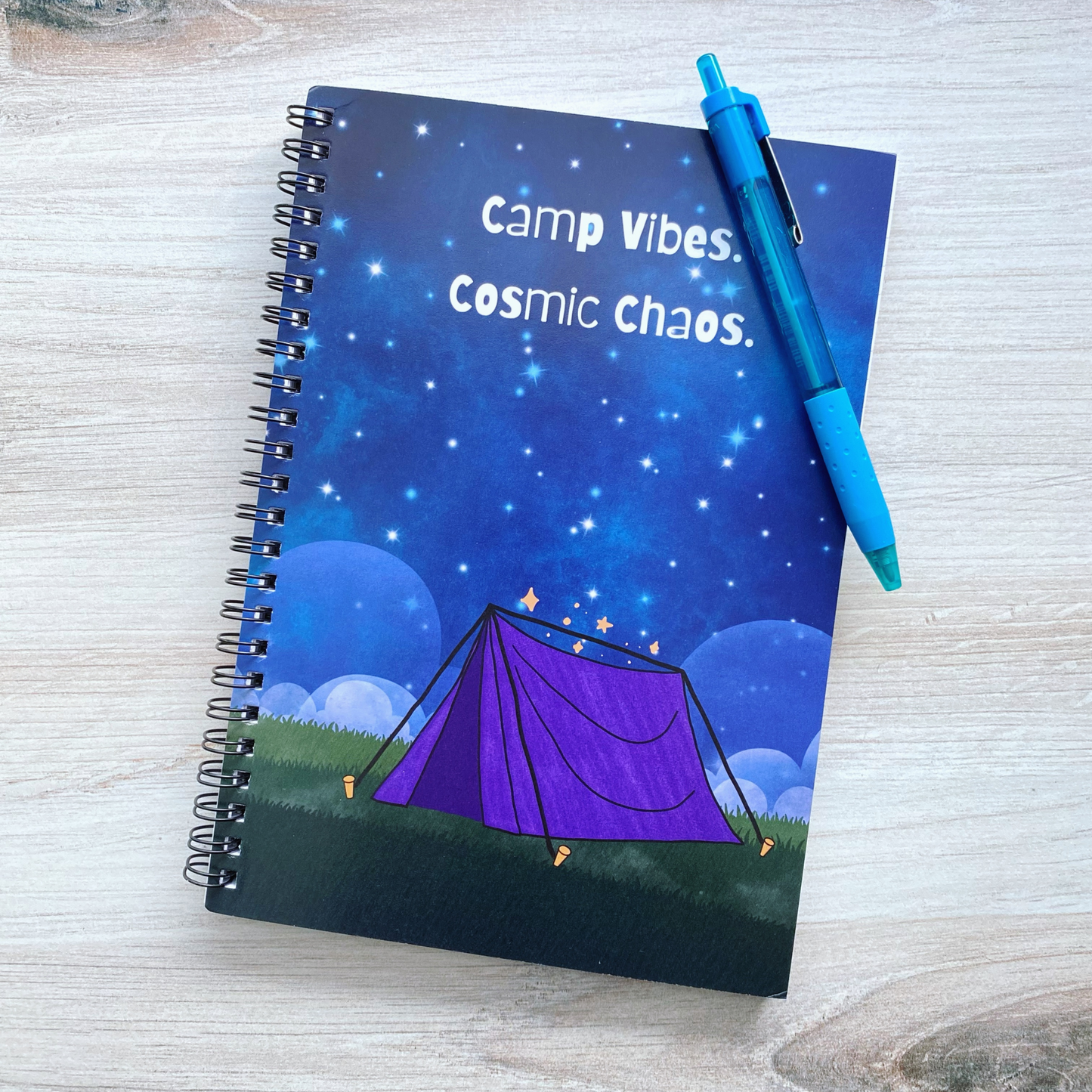 Spiral notebook cover illustrated with a purple tent under a starry night sky, reading "Camp vibes. Cosmic chaos.," resting on a wood surface next to a teal pen.