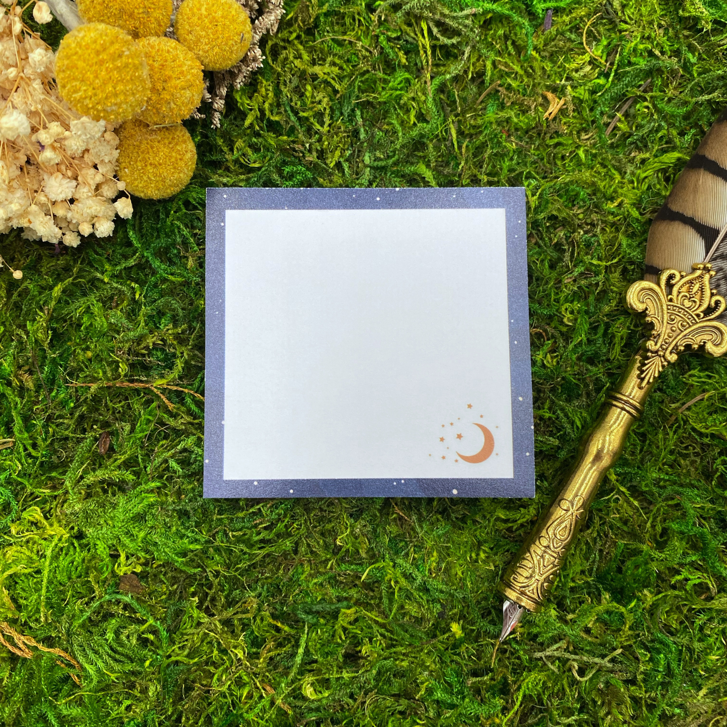 Square sticky note pad with a navy blue starfield border, a small copper crescent moon and stars in the corner, resting on moss next to dried yellow and white flowers and a gold quill pen.