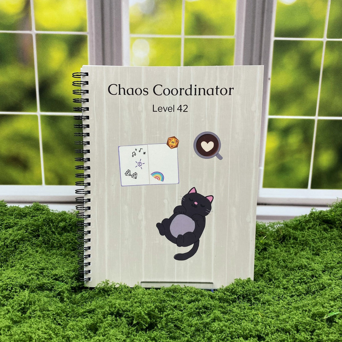 Spiral-bound wood-textured notebook reading 'Chaos Coordinator Level 42' with illustrations of a d20 die, a sleeping black cat, and a heart-shaped latte, displayed on a moss mat with a window backdrop.