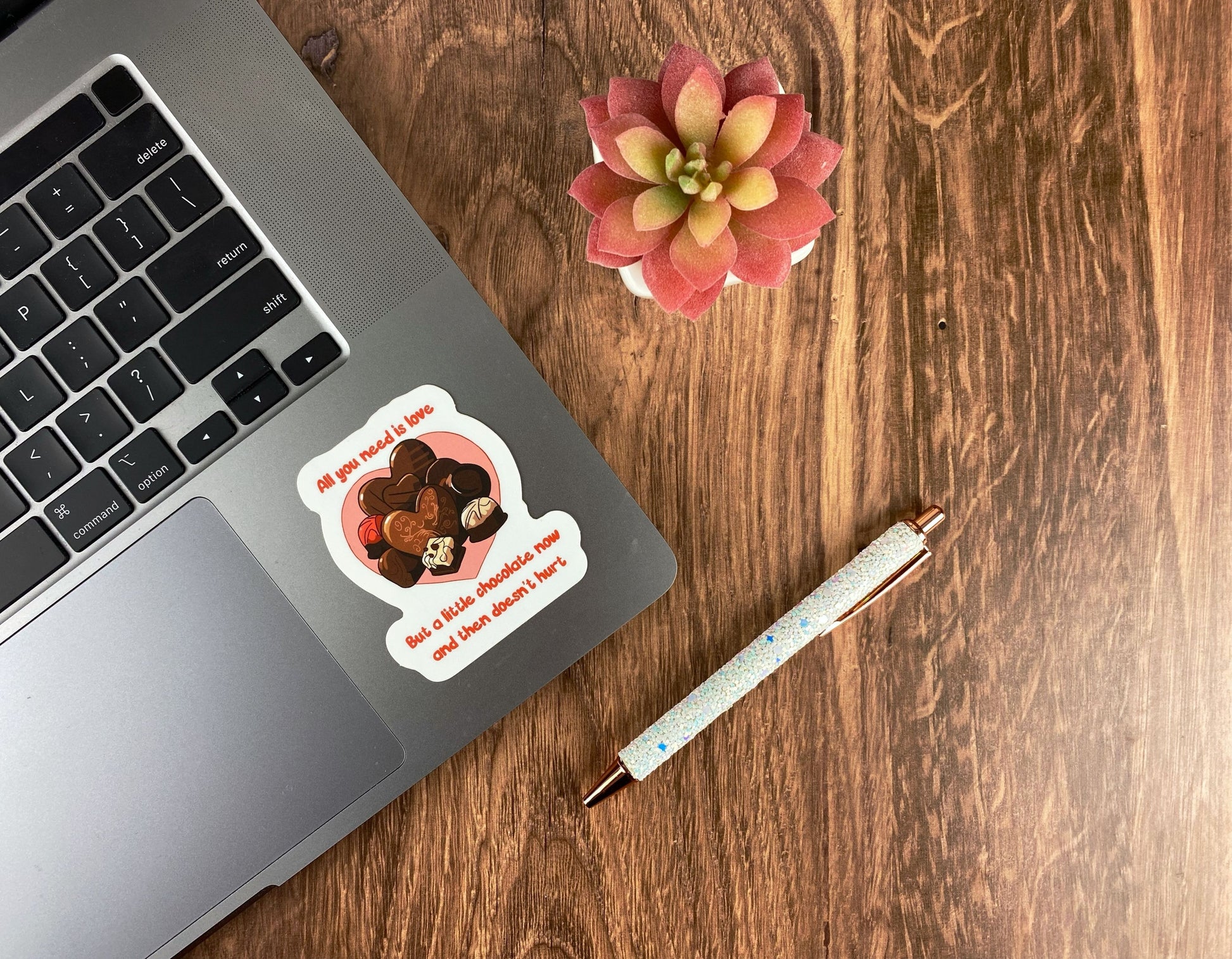 A chocolate box die-cut sticker on a laptop reading all you need is love but a little chocolate now and then doesn't hurt, next to a succulent and a glitter pen on a wood surface