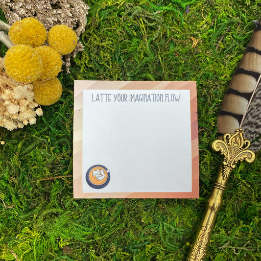 Square sticky note pad with a diagonal striped brown border, a fox illustration inside a latte cup, and the words "Latte your imagination flow," resting on moss next to dried yellow and white flowers and a gold quill pen.
