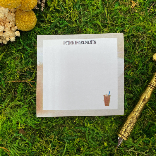 Square sticky note pad with a warm tan gradient border, a small iced coffee illustration, and the words "Potion Ingredients," resting on moss next to dried yellow and white flowers and a gold quill pen.