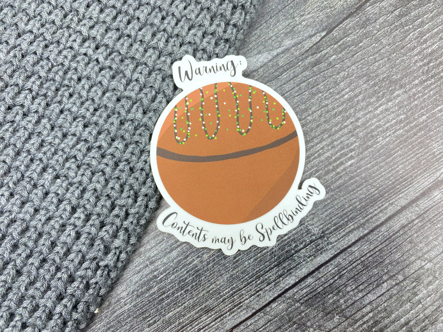 Die-cut vinyl sticker showing a drizzled hot cocoa bomb, with the text "Warning: contents may be spellbinding," on a gray knit and wood background