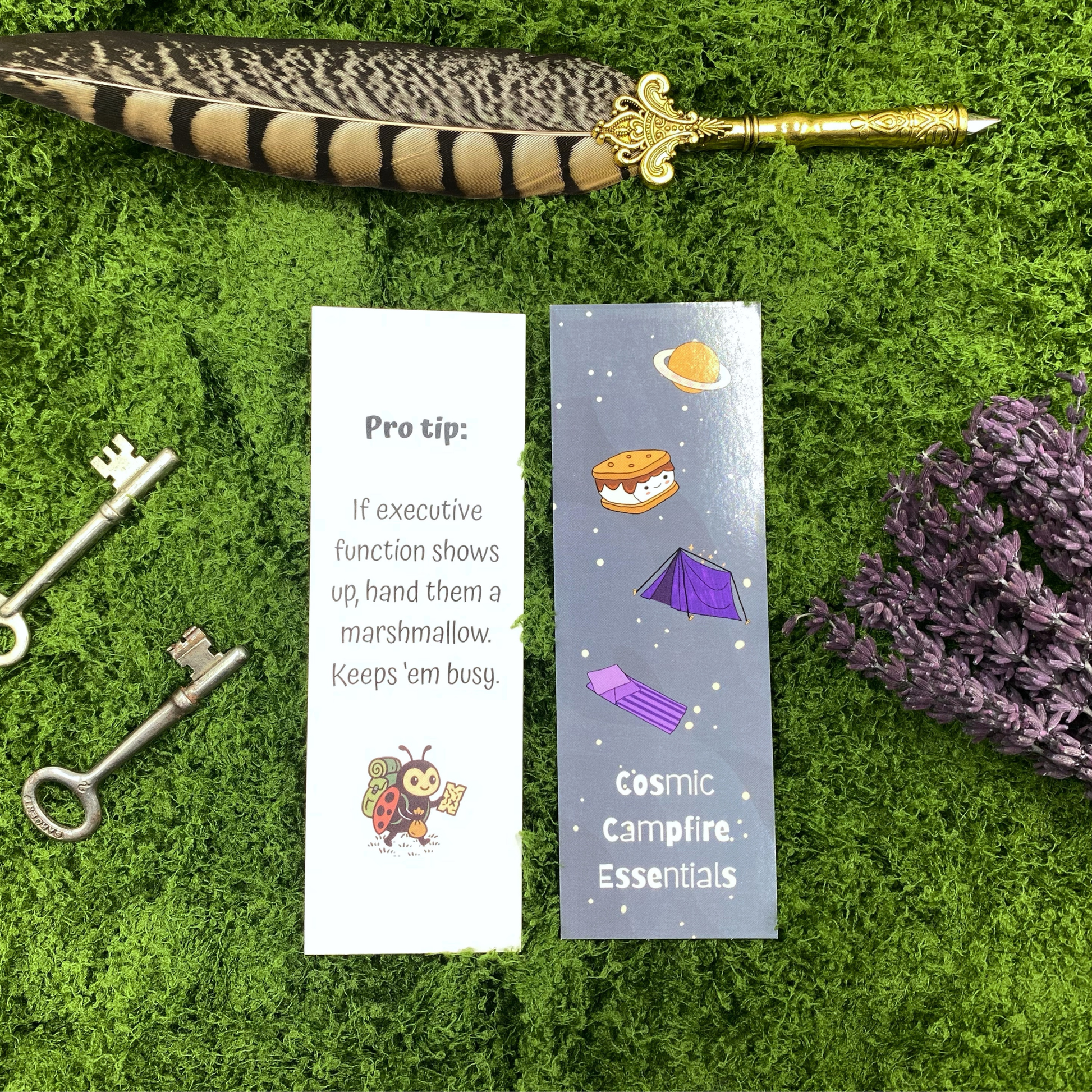 Two sides of a bookmark shown side by side, one dark purple side illustrated with a planet, s'more, tent, and sleeping bag reading "Cosmic Campfire Essentials," the other white side reading "Pro tip: If executive function shows up, hand them a marshmallow. Keeps 'em busy," with a backpacking ladybug illustration, resting on artificial grass next to two vintage keys, dried lavender, and a gold quill pen.