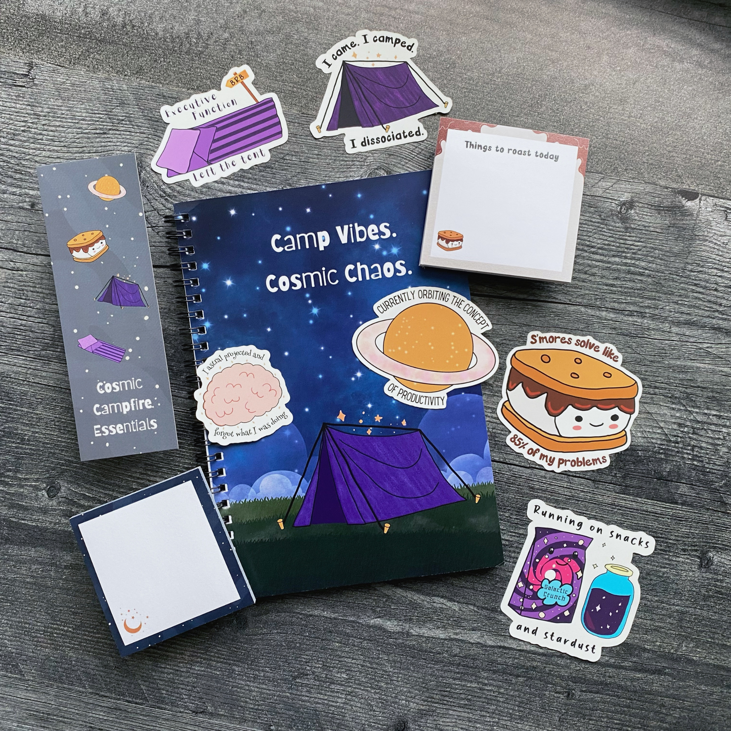 A curated gift set laid out on a gray wood background, including a starry spiral notebook reading "Camp Vibes, Cosmic Chaos," a bookmark reading "Cosmic Campfire Essentials," a sticky note pad reading "Things to Roast Today," and six camping- and space-themed stickers