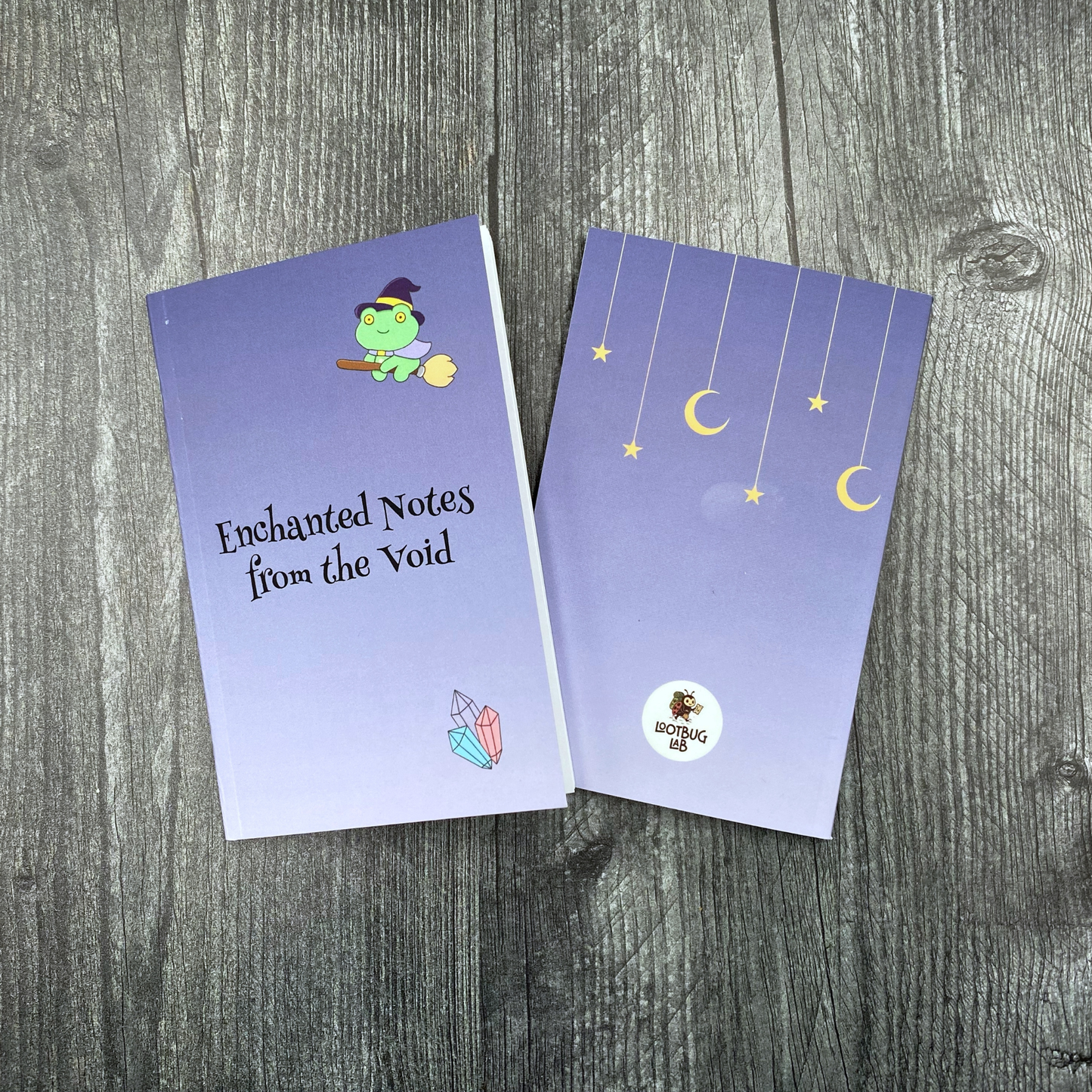Small pocket notebook cover in purple with a green frog wearing a witch hat riding a broomstick, crystals, and the words "Enchanted Notes from the Void," shown beside the back cover featuring hanging moons and stars and the Lootbug Lab logo, both on a grey wood surface.
