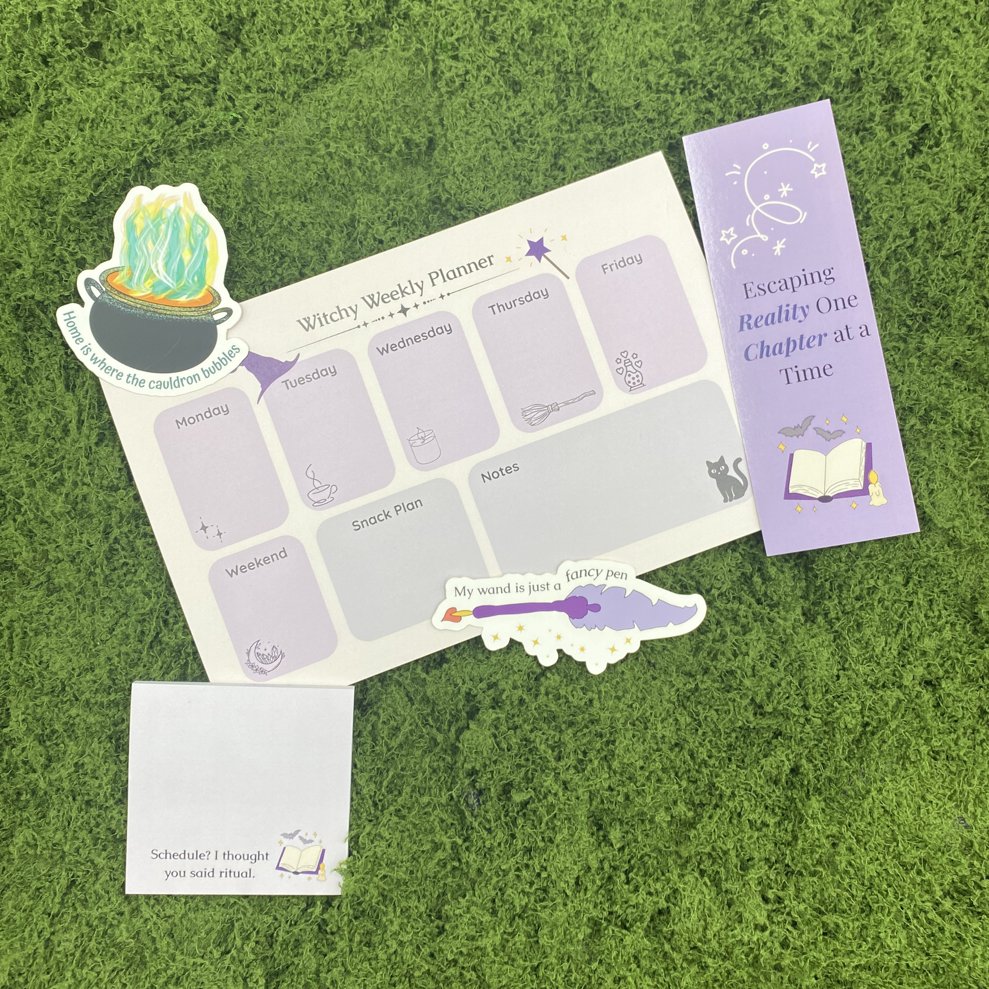 A curated gift set on artificial grass featuring a "Witchy Weekly Planner" notepad, a bubbling cauldron sticker, a quill pen sticker reading "My wand is just a fancy pen," a sticky note reading "Schedule? I thought you said ritual," and a purple bookmark reading "Escaping Reality One Chapter at a Time."