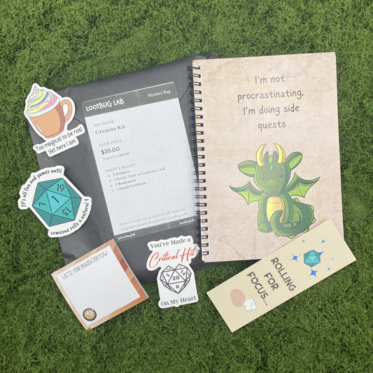 A Lootbug Lab creative kit mystery bag with a reveal card surrounded by sample items including stickers, a bookmark, a small notebook, and a sticky note pad on a green moss surface