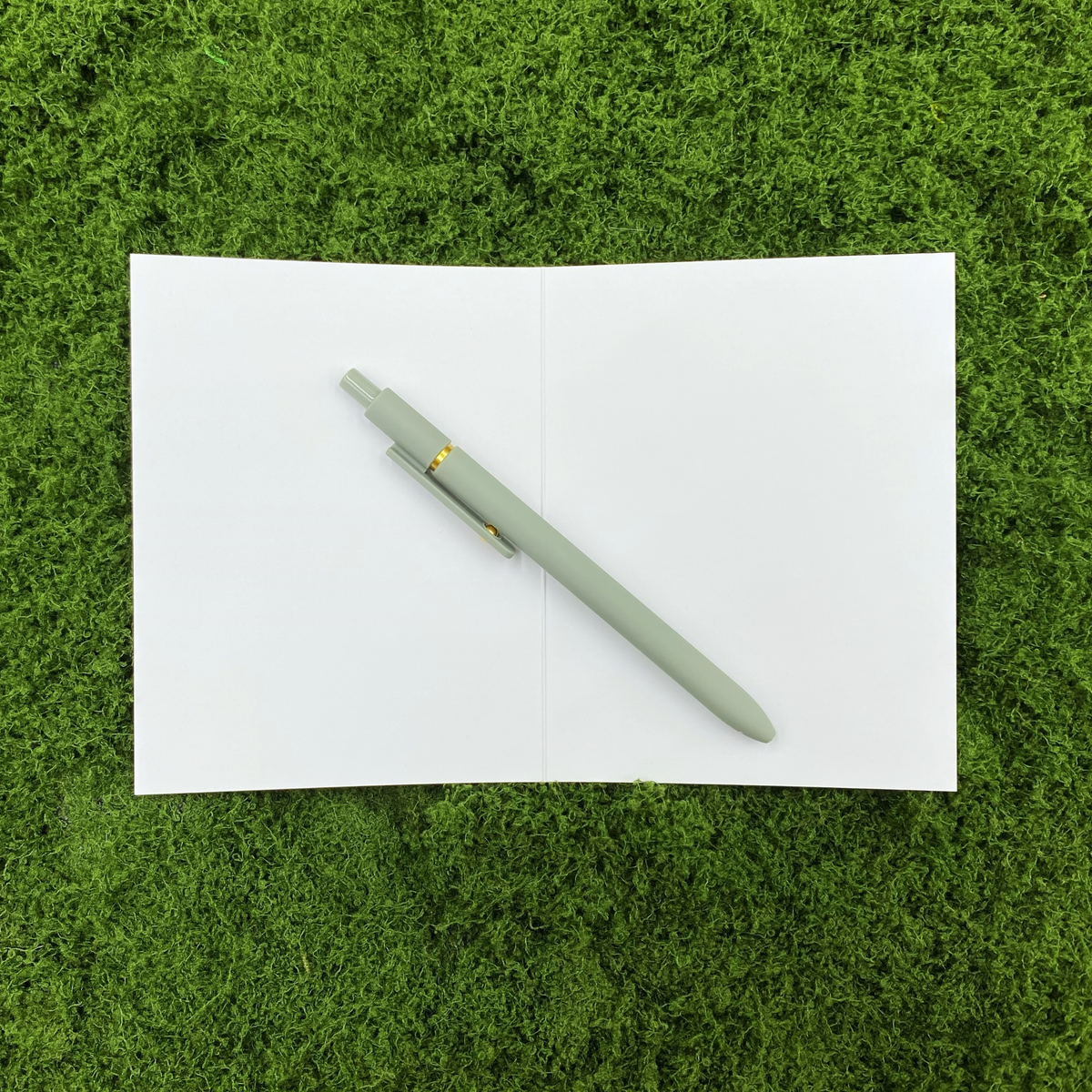 Card opened to show the blank white interior, ready for a handwritten message, with a sage green pen laid across the crease on a mossy background.