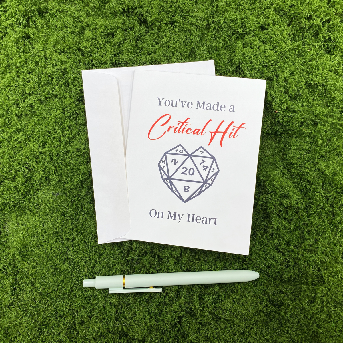 Folded greeting card reading "You've Made a Critical Hit On My Heart" with a heart-shaped d20 illustration, resting on a white envelope on a mossy surface, with a sage green pen below.