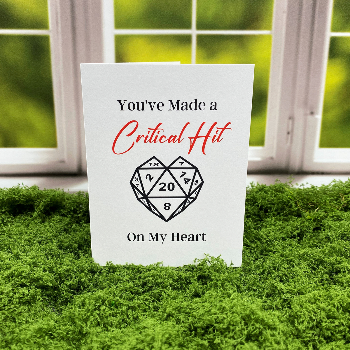 Greeting card that reads "You've Made a Critical Hit On My Heart," with "Critical Hit" in red script, above a black-and-white heart-shaped twenty-sided die showing the number 20, standing upright on a moss surface in front of a white-framed window.