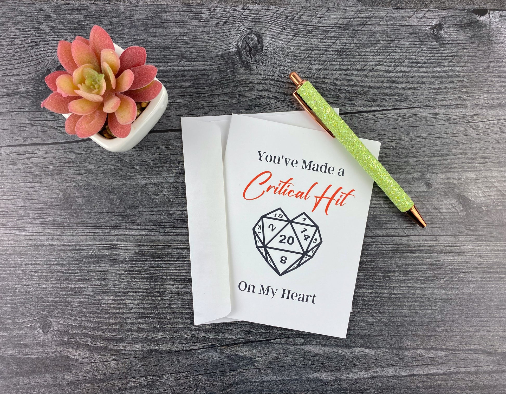 Greeting card and matching envelope, front reading "You've made a critical hit on my heart" with a heart-shaped d20 die illustration, resting on a grey wood surface next to a green glitter pen and small succulent.