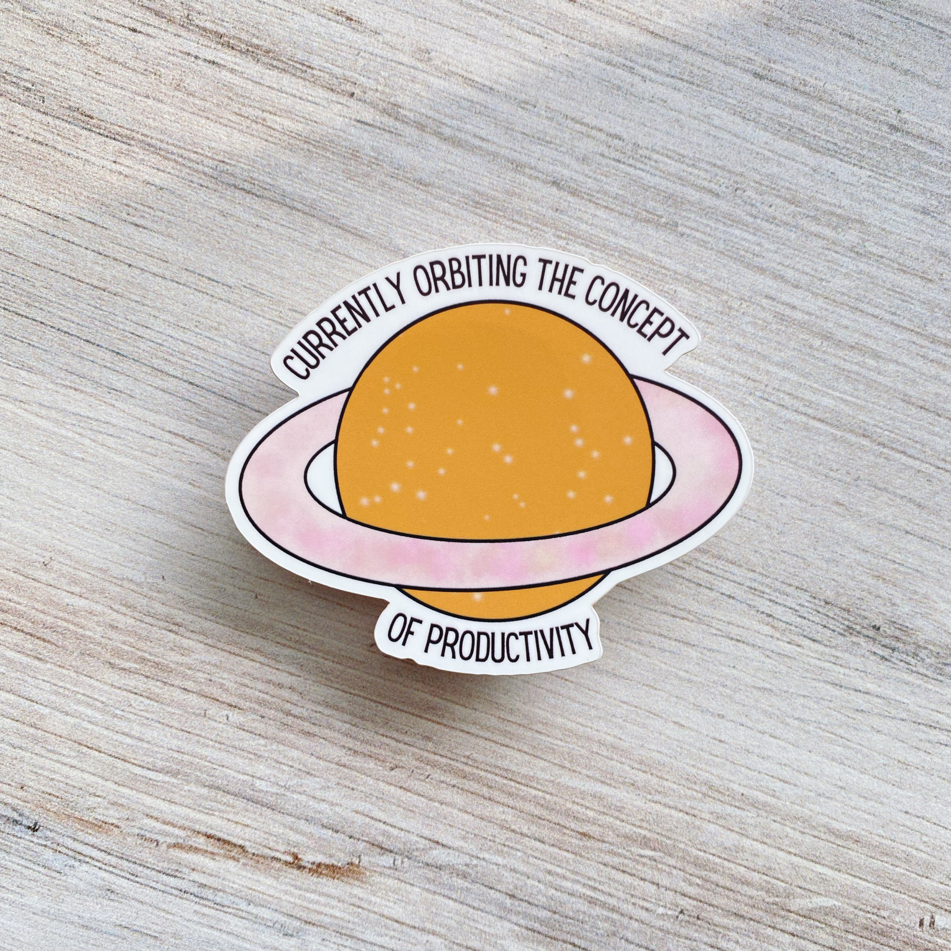 Die-cut vinyl sticker showing a starry orange planet with pink rings, with the text "Currently orbiting the concept of productivity," on a light wood background