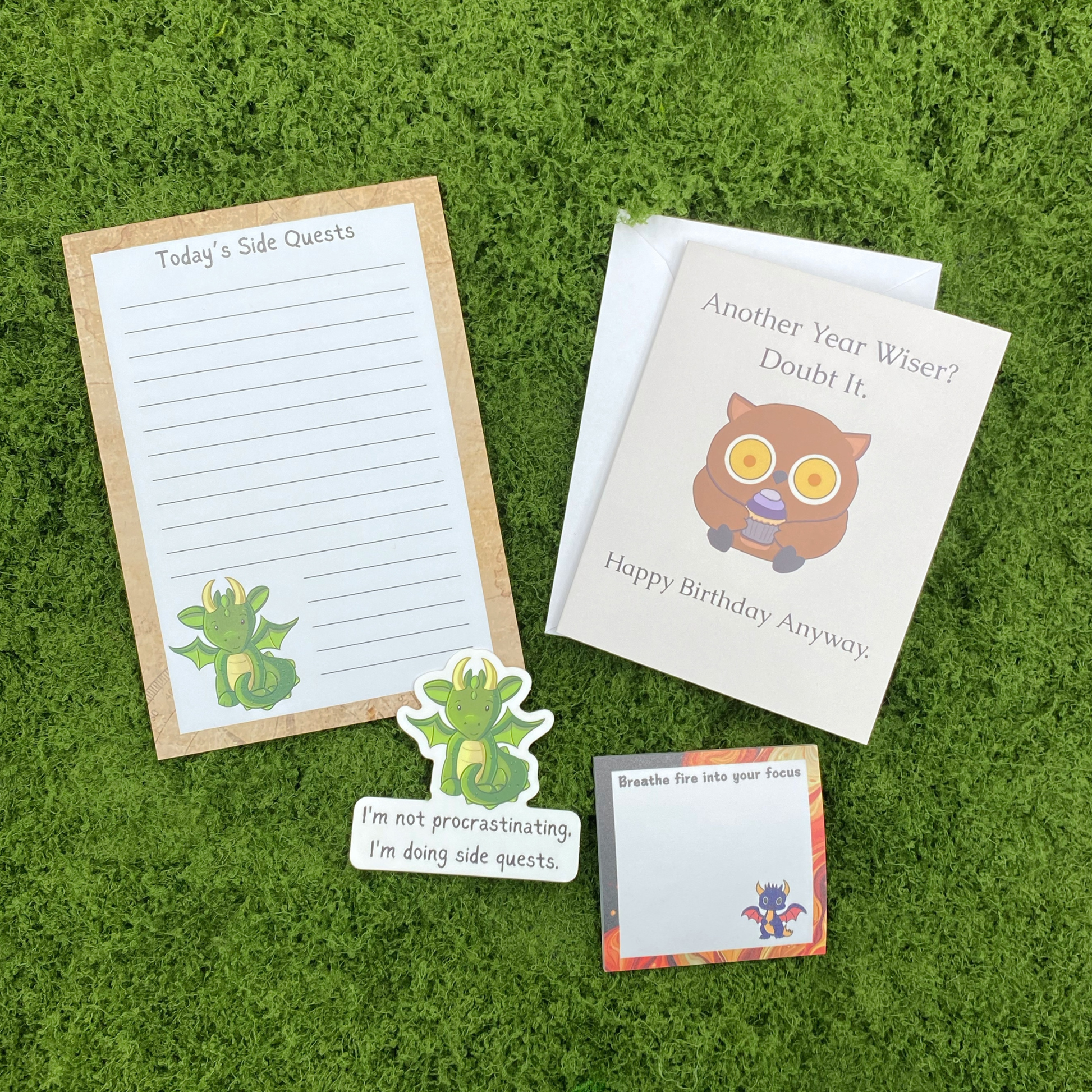 A curated gift set on artificial grass featuring a "Today's Side Quests" notepad with a dragon illustration, a dragon sticker, a "Breathe Fire Into Your Focus" sticky note, and a greeting card that reads "Another Year Wiser? Doubt It." with an owl holding a cupcake.