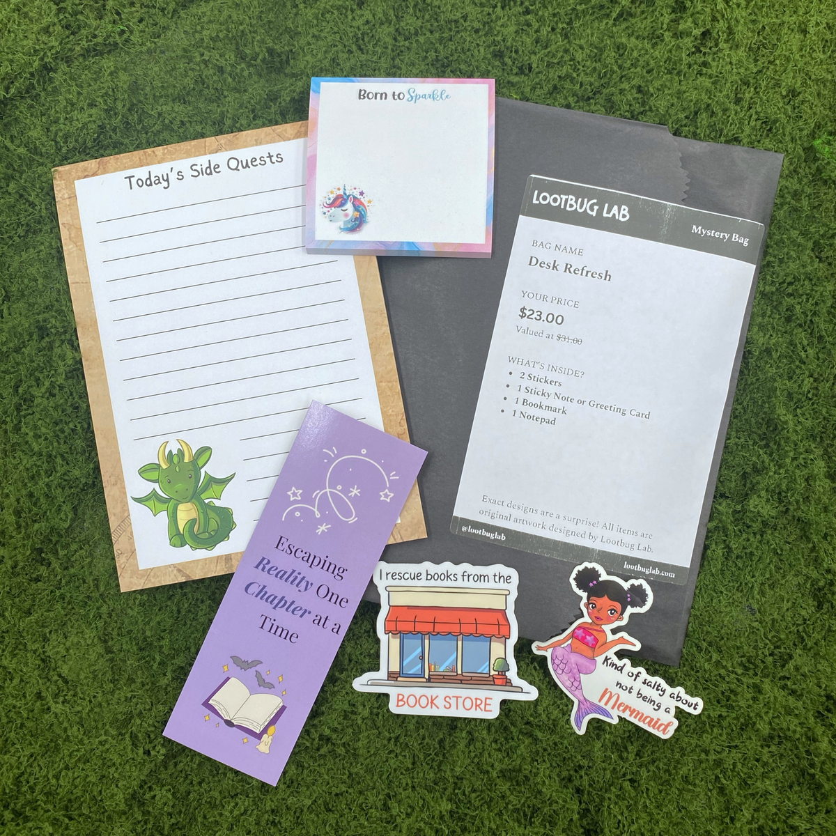 A Lootbug Lab "Desk Refresh" mystery bag price tag listing $23.00 (valued at $31.00) and contents of 2 stickers, 1 sticky note or greeting card, 1 bookmark, and 1 notepad, on a grey paper bag surrounded by example items: a "Today's Side Quests" notepad with a dragon sticker, a "Born to Sparkle" unicorn sticky note, a bookmark reading "Escaping Reality One Chapter at a Time," a bookstore sticker, and a mermaid sticker, on artificial grass.