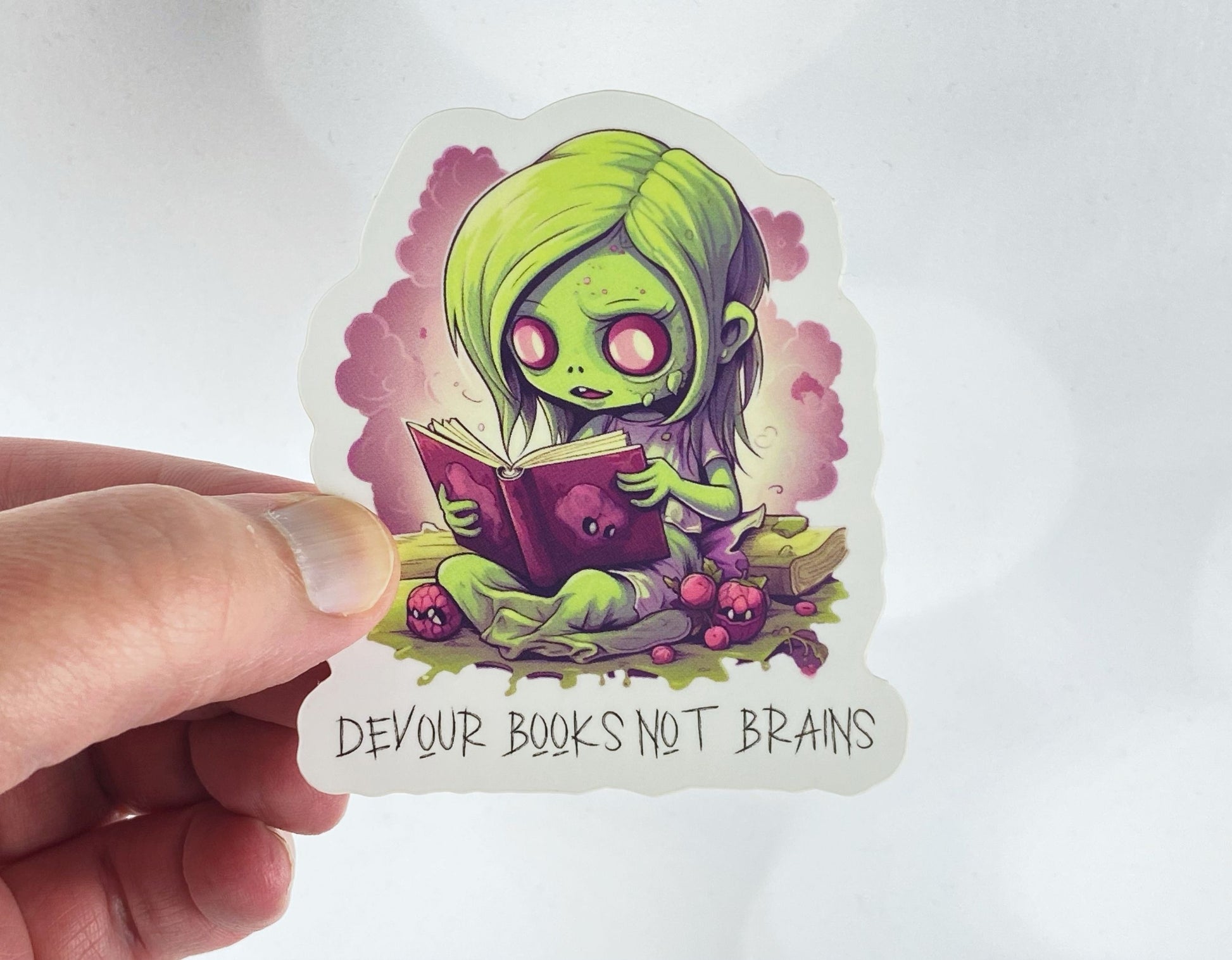 A hand holds a sticker illustration of a green zombie girl reading a book, reading "Devour books not brains," against a white background.