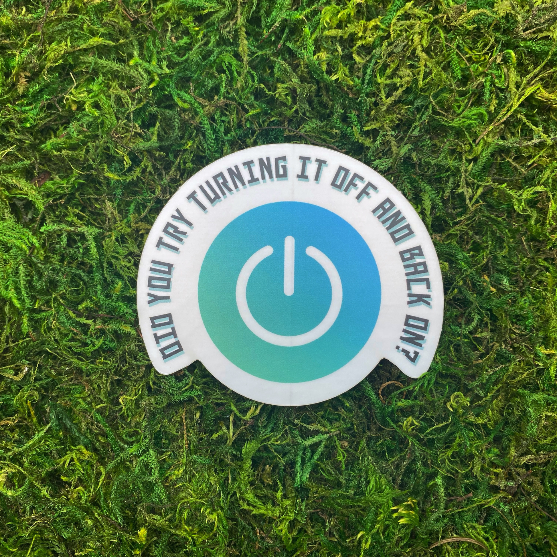 A round sticker with a teal power button icon and the text "Did you try turning it off and back on?" resting on artificial moss.