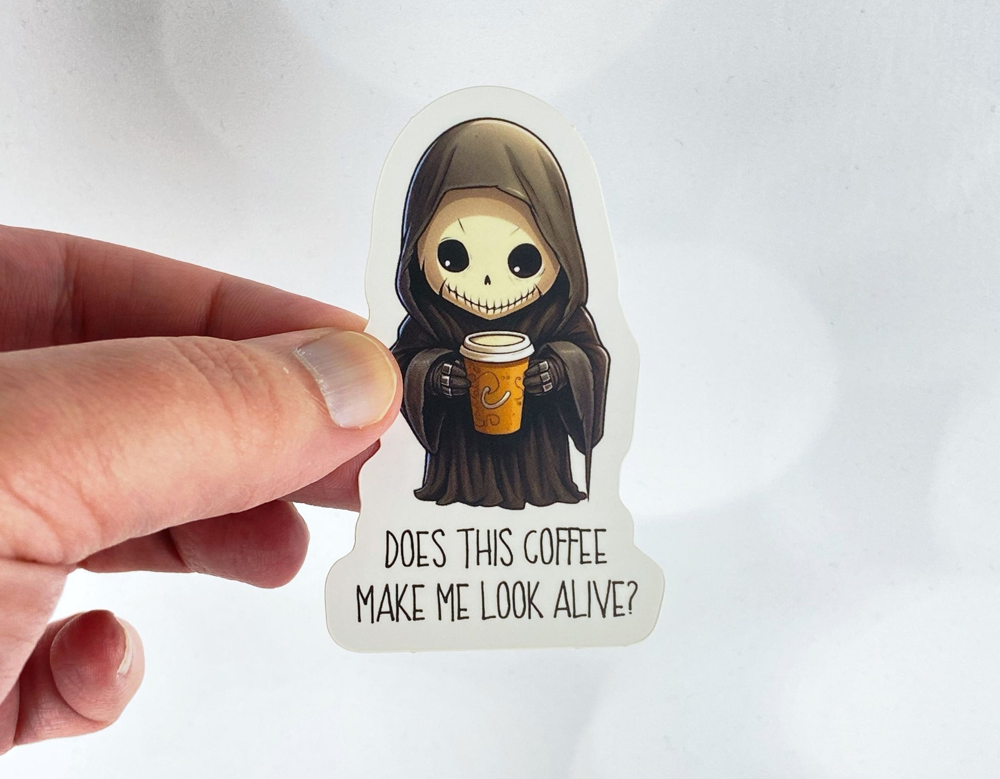 A hand holds a sticker illustration of a small hooded grim reaper holding a coffee cup, reading "Does this coffee make me look alive?" against a white background.