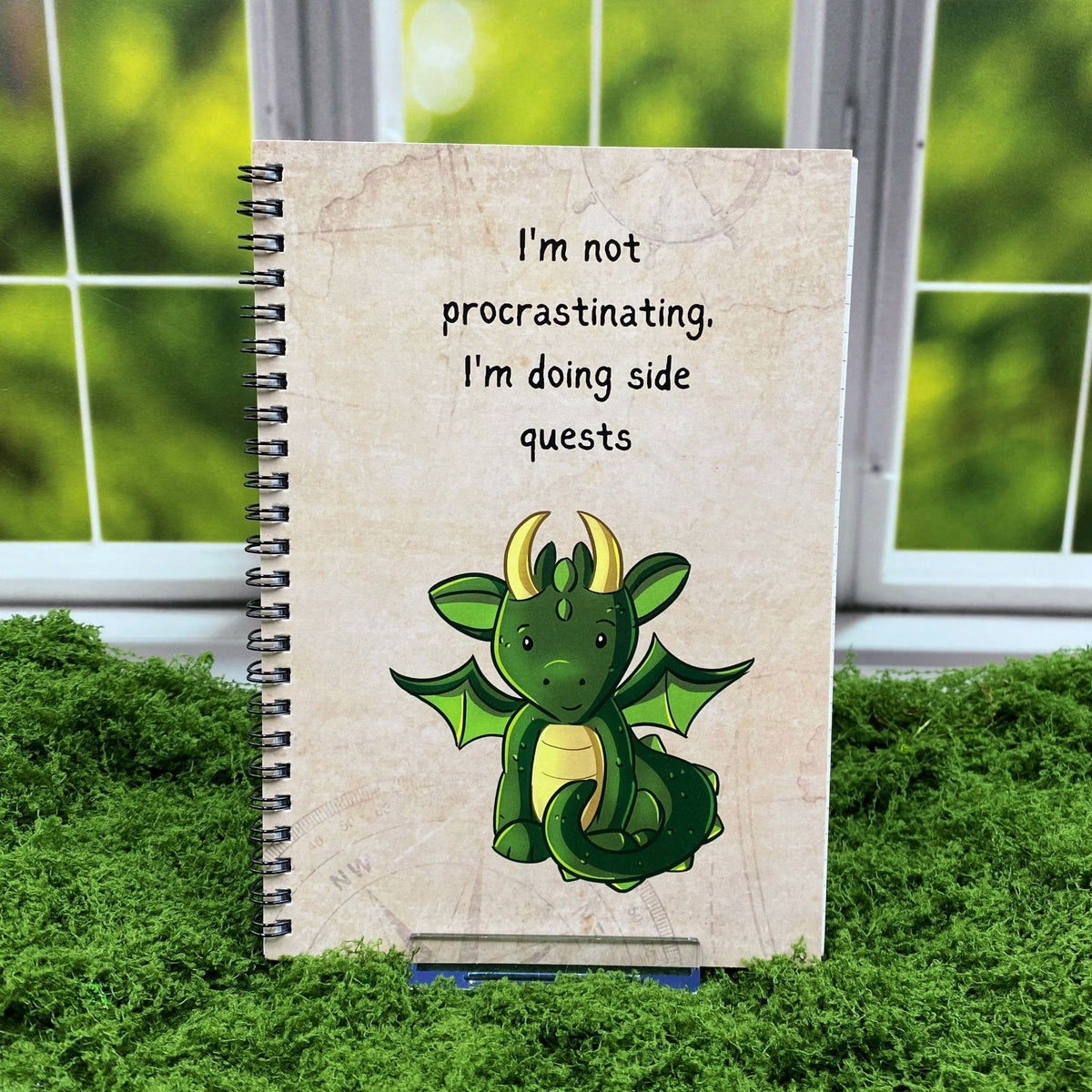 Spiral-bound notebook with a tan map-textured cover reading 'I'm not procrastinating. I'm doing side quests' above a cute green dragon illustration, displayed on a moss mat with a window backdrop.