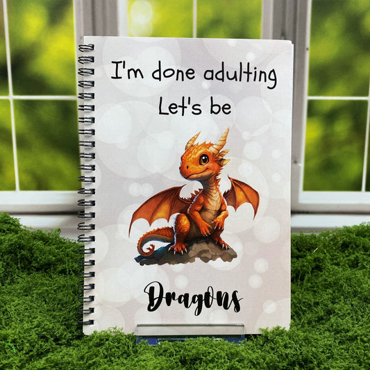 Spiral-bound notebook with a pale bokeh-patterned cover featuring a baby orange dragon and text reading 'I'm done adulting. Let's be Dragons,' displayed on a moss mat with a window backdrop.