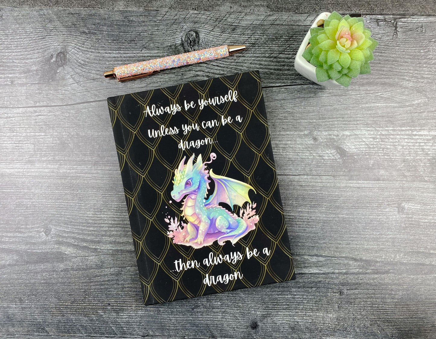 Black hardcover journal with a gold scale pattern, an illustrated pastel baby dragon, and the words "Always be yourself, unless you can be a dragon, then always be a dragon," resting on a grey wood surface next to a glitter pen and small succulent.