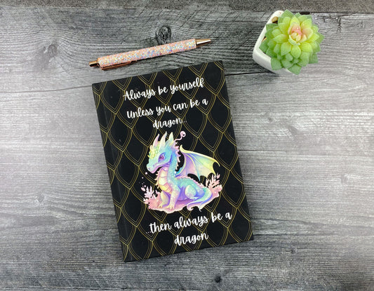 Black hardcover journal with a gold scale pattern, an illustrated pastel baby dragon, and the words "Always be yourself, unless you can be a dragon, then always be a dragon," resting on a grey wood surface next to a glitter pen and small succulent.