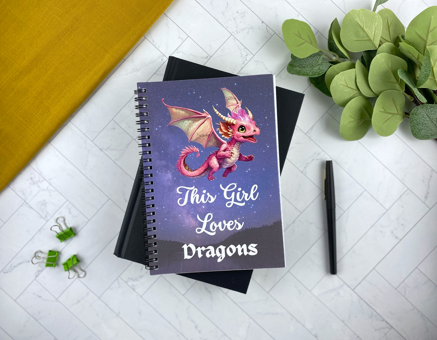 Spiral notebook cover with a starry night sky, a pink baby dragon with folded wings, and the words "This Girl Loves Dragons," resting on a black notebook with green binder clips, a black pen, and eucalyptus greenery nearby.
