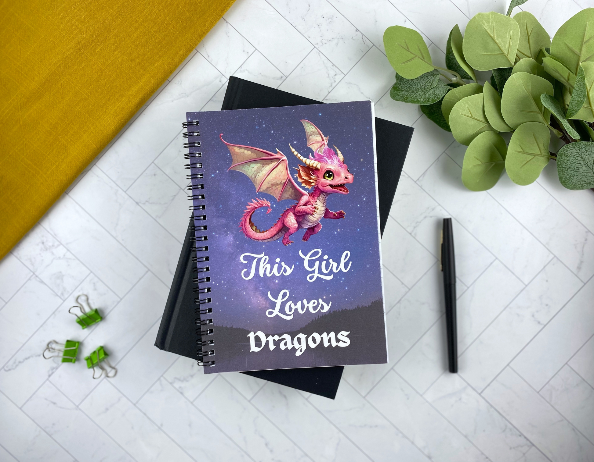 Spiral notebook cover with a starry night sky, a pink baby dragon with folded wings, and the words "This Girl Loves Dragons," resting on a black notebook with green binder clips, a black pen, and eucalyptus greenery nearby.
