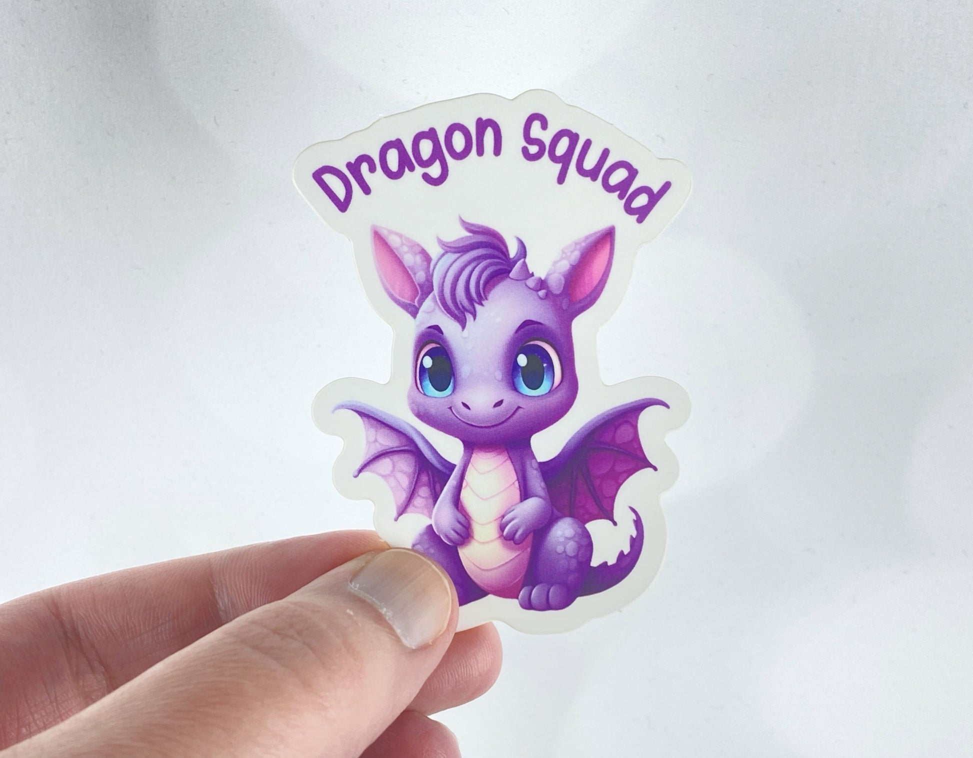 A hand holds a sticker illustration of a wide-eyed purple baby dragon with folded wings, reading "Dragon Squad," against a white background.