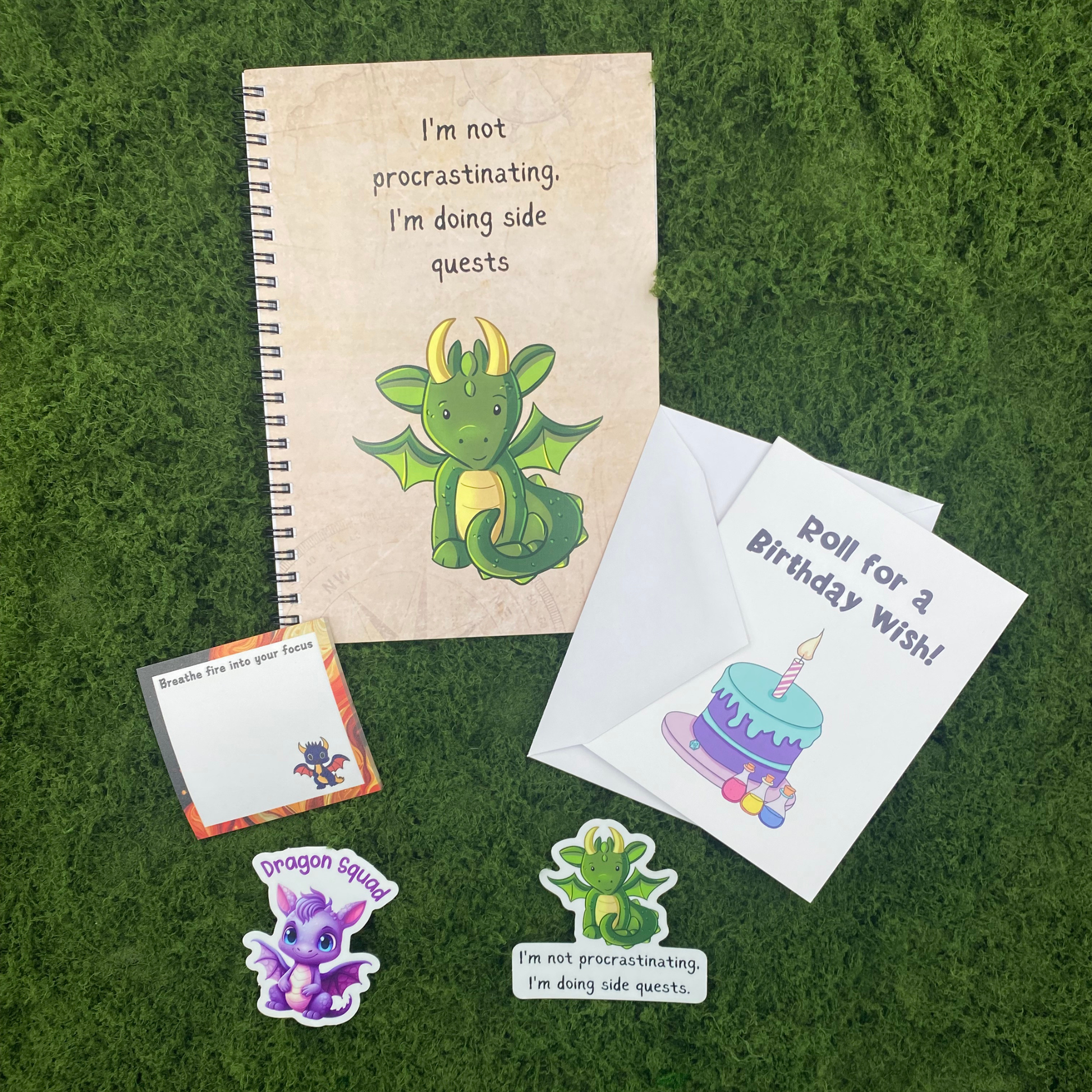 A birthday gift set including an I'm not procrastinating I'm doing side quests spiral notebook, a breathe fire into your focus sticky note pad, a roll for a birthday wish greeting card with envelope, and two dragon die-cut stickers, arranged on a green moss surface