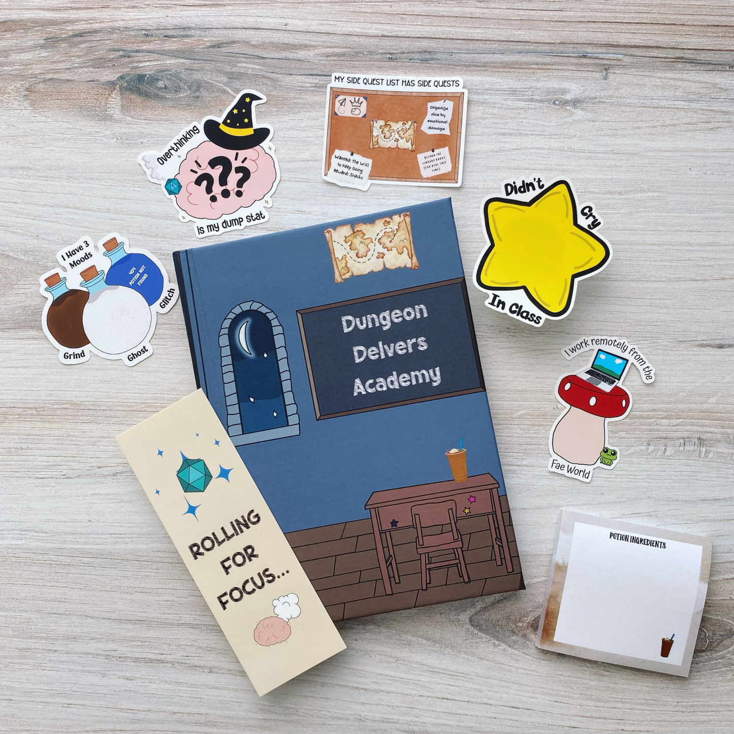 A curated gift set laid out on a light wood background, including a spiral notebook reading "Dungeon Delvers Academy," a bookmark reading "Rolling for Focus," a sticky note pad reading "Potion Ingredients," and five tabletop RPG and school-humor stickers