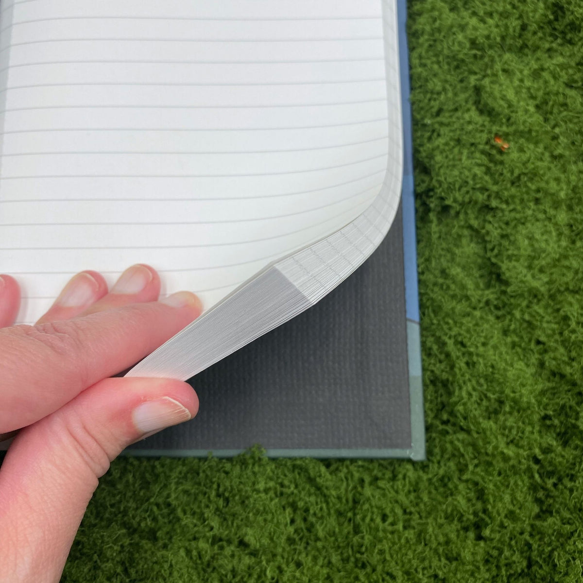 Close-up of a hand fanning through the lined pages of the journal, showing the plain paper lining inside the hardcover binding.