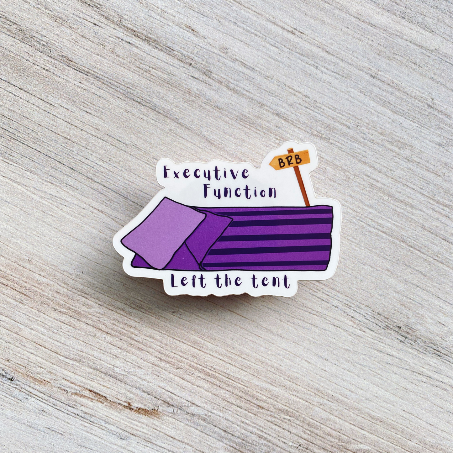Die-cut vinyl sticker showing a purple sleeping bag with a "BRB" sign on a post, with the text "Executive function left the tent," on a light wood background