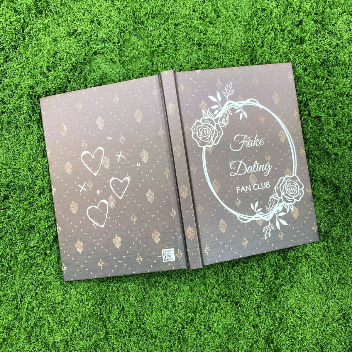 Open brown hardcover journal showing hand-drawn hearts and doodles on the back cover and the rose wreath 'Fake Dating Fan Club' design on the front.