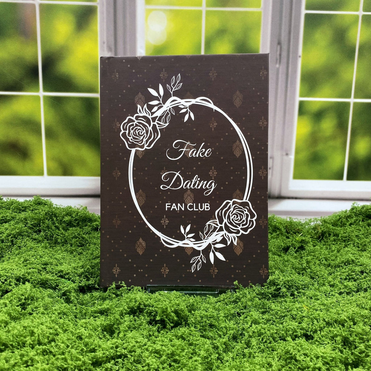 Brown hardcover journal with a damask pattern and a rose wreath frame around text reading 'Fake Dating Fan Club,' displayed on a moss mat with a window backdrop.