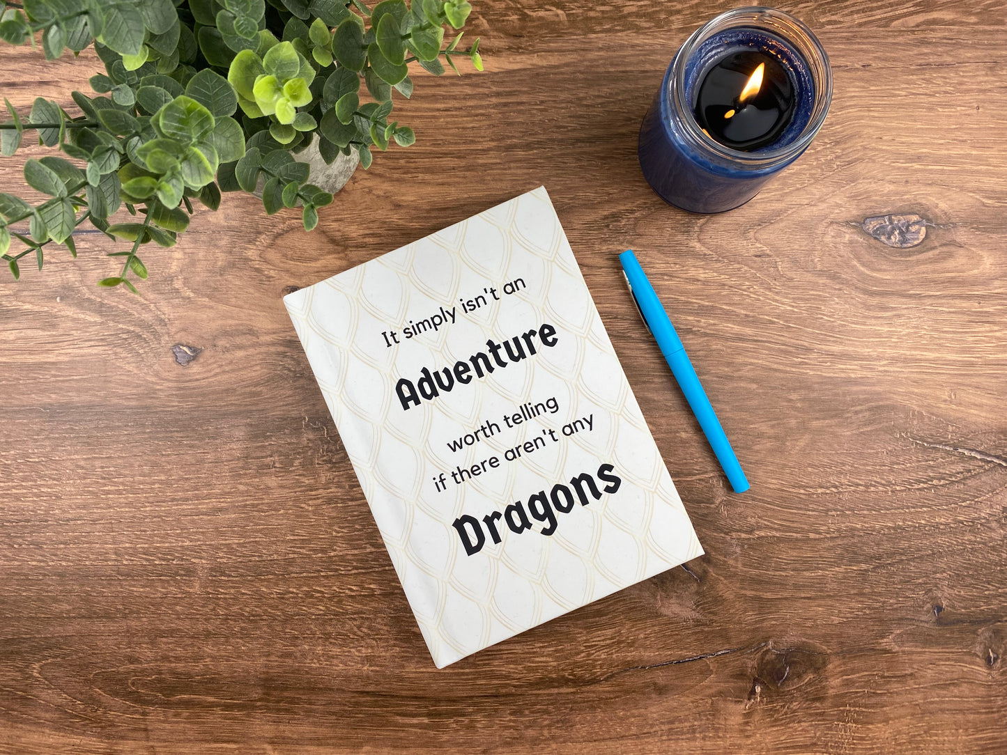 Hardcover journal with a cream dragon-scale pattern and the words "It simply isn't an adventure worth telling if there aren't any dragons," resting on a wood surface next to a lit candle and a blue pen.
