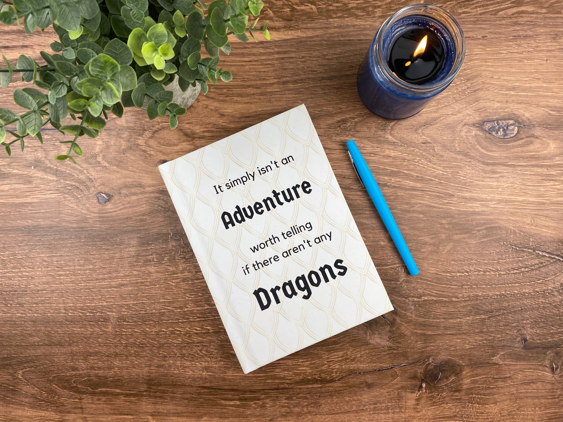 Hardcover journal with a cream dragon-scale pattern and the words "It simply isn't an adventure worth telling if there aren't any dragons," resting on a wood surface next to a lit candle and a blue pen.