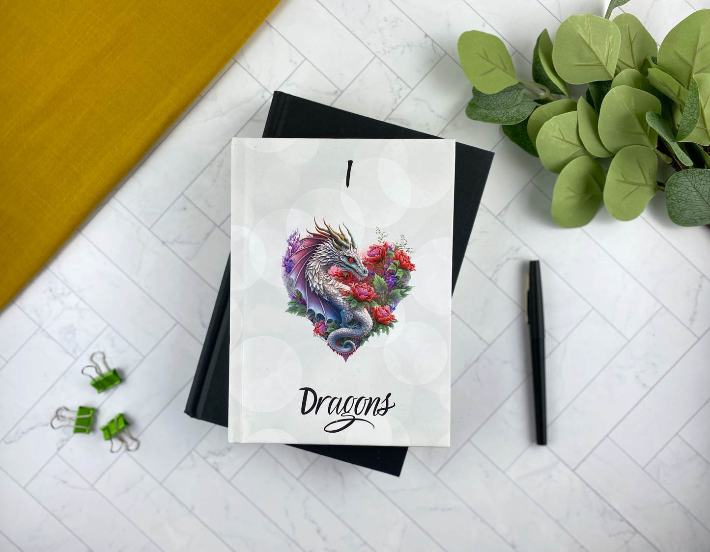 Hardcover journal with a soft bokeh background, an illustrated silver dragon curled into a heart shape surrounded by red and purple flowers, reading "I [heart] Dragons," resting on a black notebook with green binder clips and a black pen nearby.