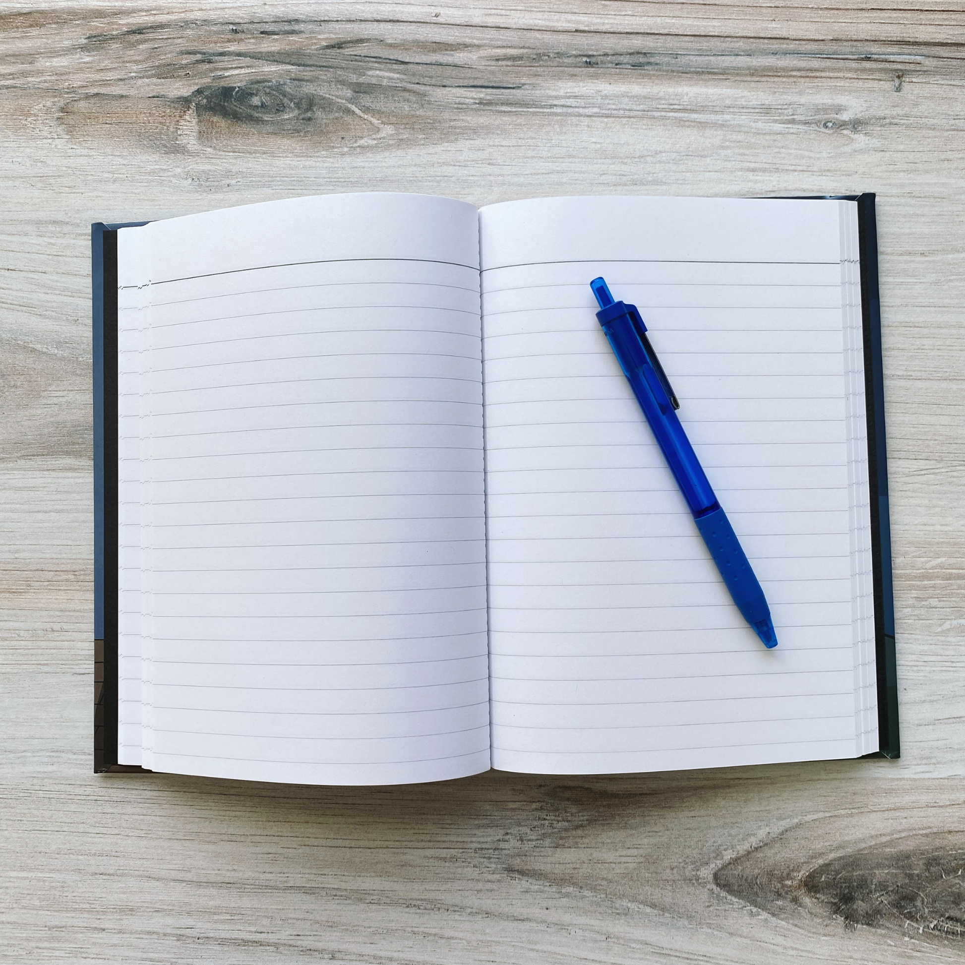 Open journal showing blank lined pages with a blue pen resting across them, on a wood surface.