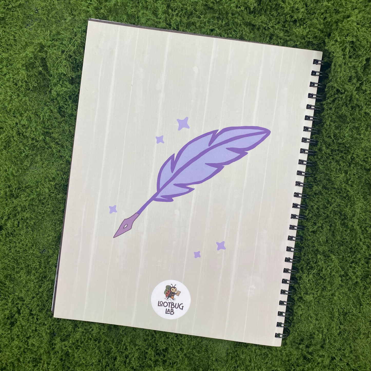 Notebook back cover with a purple feather quill and stars on a wood-textured background, with the Lootbug Lab logo, on artificial grass.