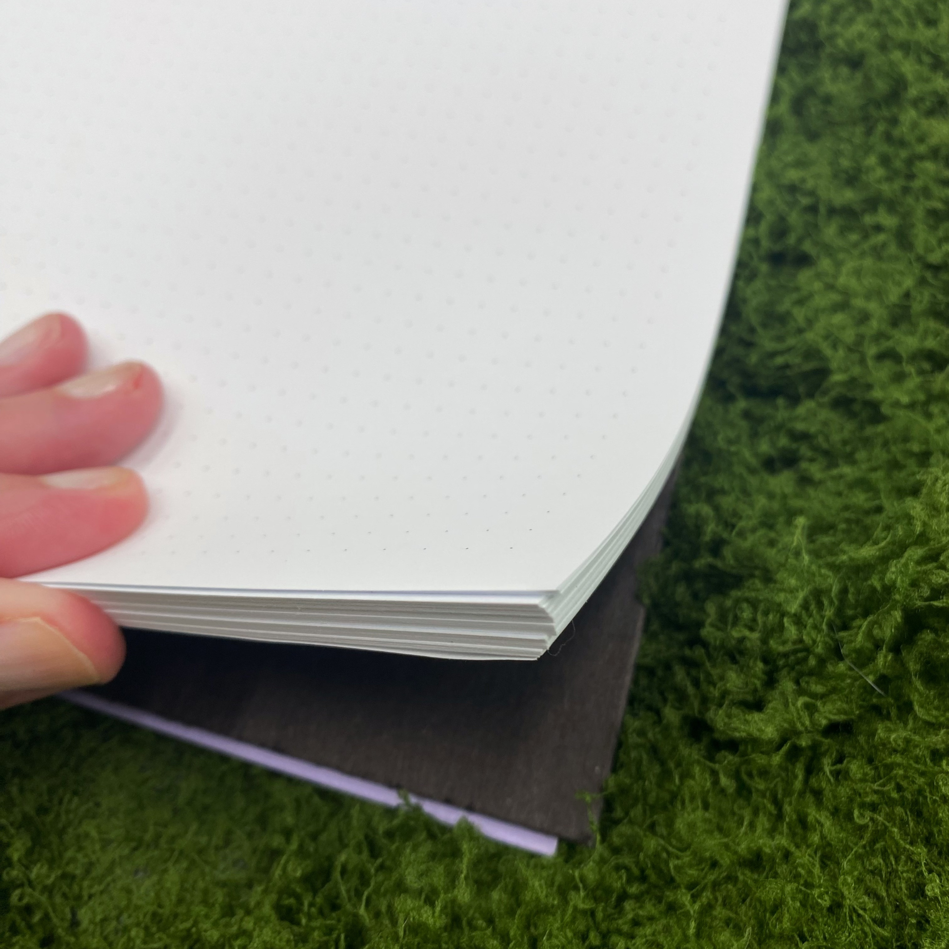 Close-up of dot grid pages inside a notebook with a lavender-edged cover peeking out, shown on an artificial grass surface.