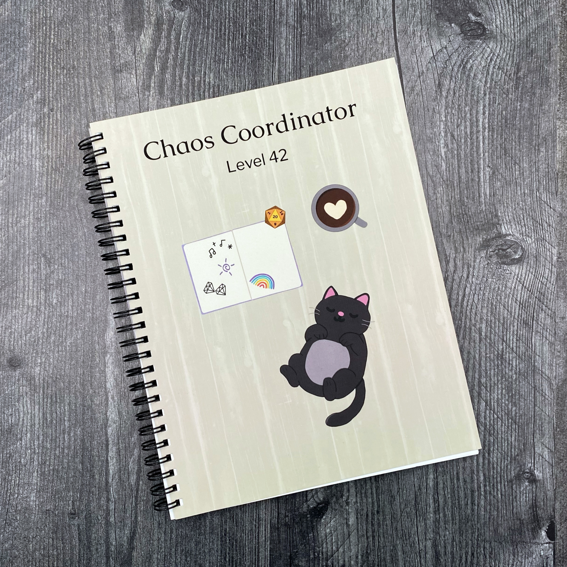 Notebook cover reading "Chaos Coordinator Level 42" with a sleepy black cat, a coffee cup with a heart in the foam, an open notebook with doodles, and a d20 die, on a grey wood surface.